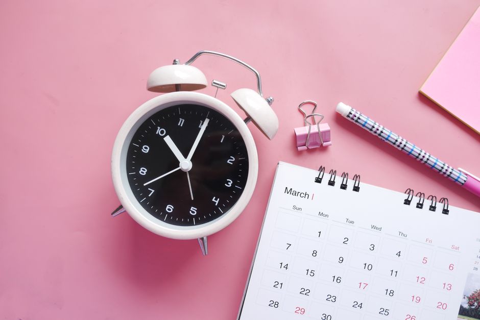 Flat lay of a pink-themed workspace featuring an alarm clock, calendar, pen, and paper clip on a pastel surface.